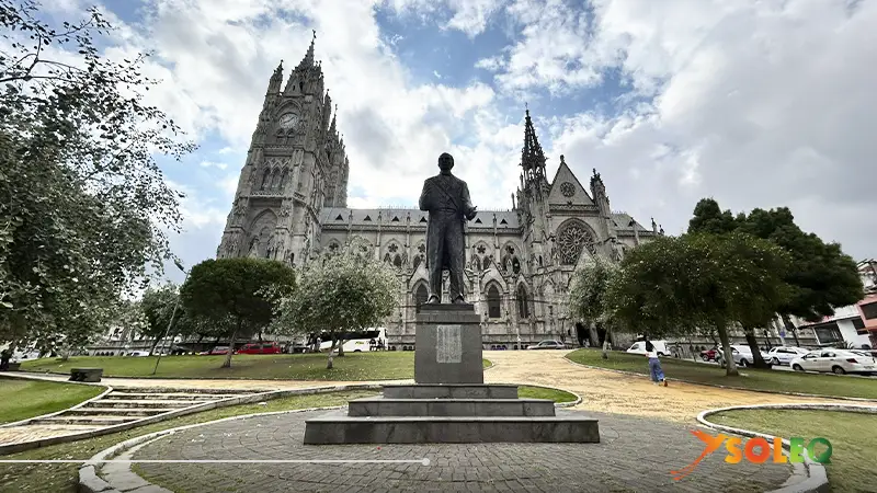 Vista frontal de la Basílica del Voto Nacional en Quito, con arquitectura neogótica y un monumento en primer plano.