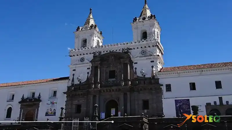 Fachada de la Iglesia de San Francisco en el Centro Histórico de Quito, con sus icónicas torres gemelas blancas y su entrada barroca bajo un cielo azul despejado.