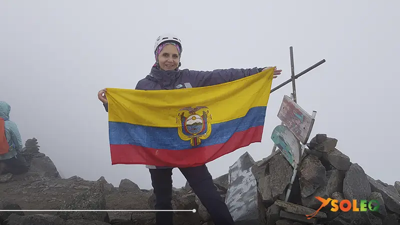 Smiling hiker on the summit of Volcán Corazón in Ecuador, proudly holding the Ecuadorian flag with the Andes in the background.