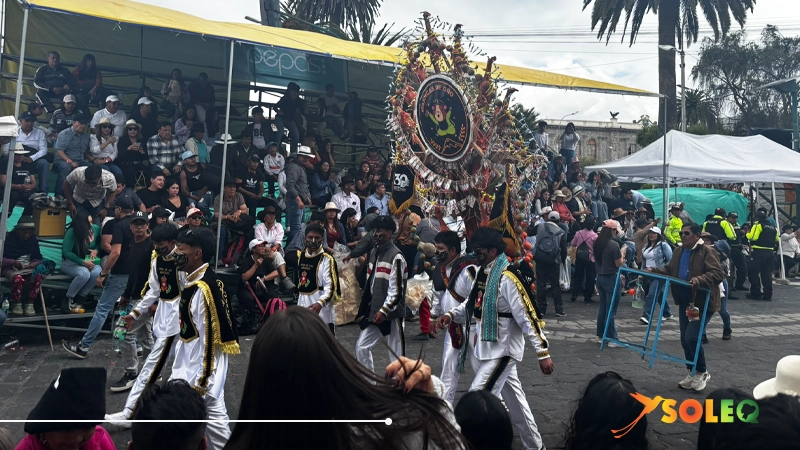 Traditional dancers in ornate costumes participating in the Mama Negra festival parade in Latacunga, Ecuador, with spectators watching from grandstands.