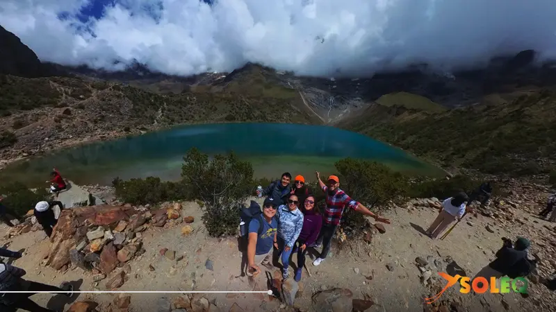 Laguna Humantay en Perú, con aguas turquesas y montañas nubladas al fondo; grupo de viajeros en primer plano en un mirador rocoso.