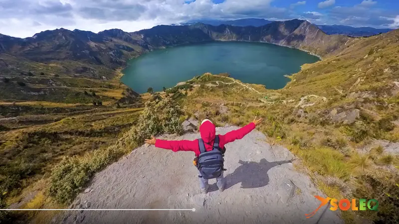 Un excursionista con los brazos abiertos contemplando el lago turquesa del cráter Quilotoa desde un pico rocoso durante la ruta de senderismo Quilotoa Loop en Ecuador.