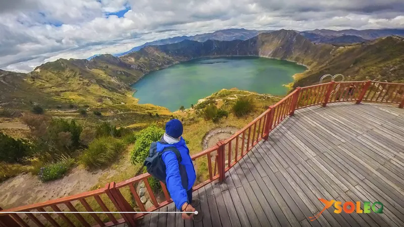 Un excursionista con chaqueta azul contempla el lago del cráter Quilotoa desde el mirador de madera Shalala durante la ruta de senderismo Quilotoa Loop en Ecuador.