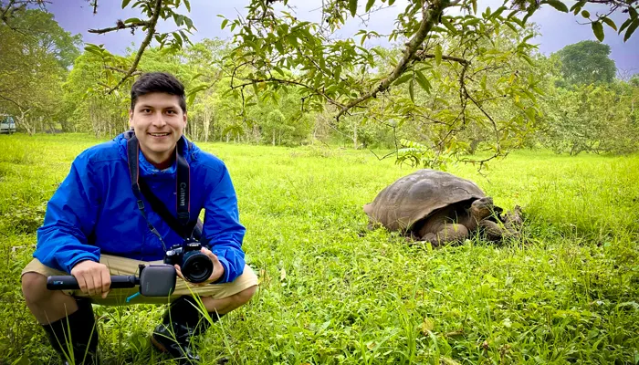 Guía naturalista arrodillado sobre el césped en Galápagos sosteniendo una cámara junto a una tortuga gigante en un paisaje verde.