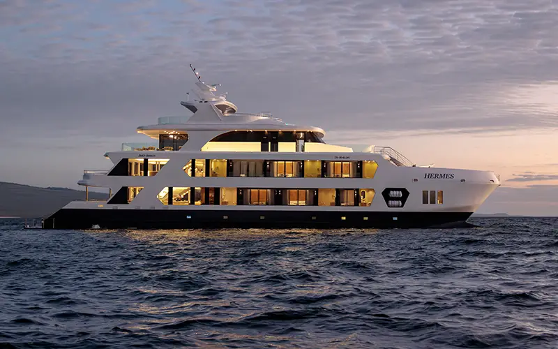 Luxury expedition yacht cruising in the Galápagos Islands at sunset with illuminated cabins.