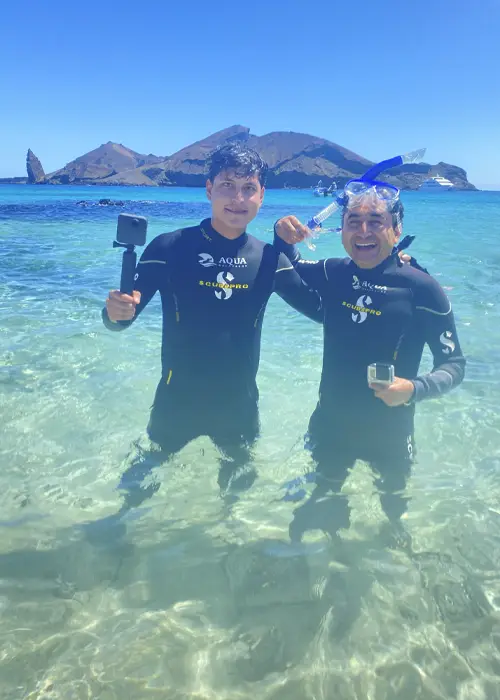 Dos hombres con trajes de neopreno de pie en aguas turquesas poco profundas en las Galápagos, sosteniendo cámaras de acción y equipo de snorkel con islas volcánicas de fondo.