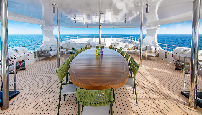 Cubierta soleada de un crucero de lujo en Galápagos con una elegante mesa de comedor de madera, asientos lounge modernos y vistas panorámicas al océano bajo un cielo azul despejado.